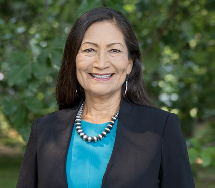 Portrait of Secretary Deb Haaland wearing a blue dress, smiling in front of trees