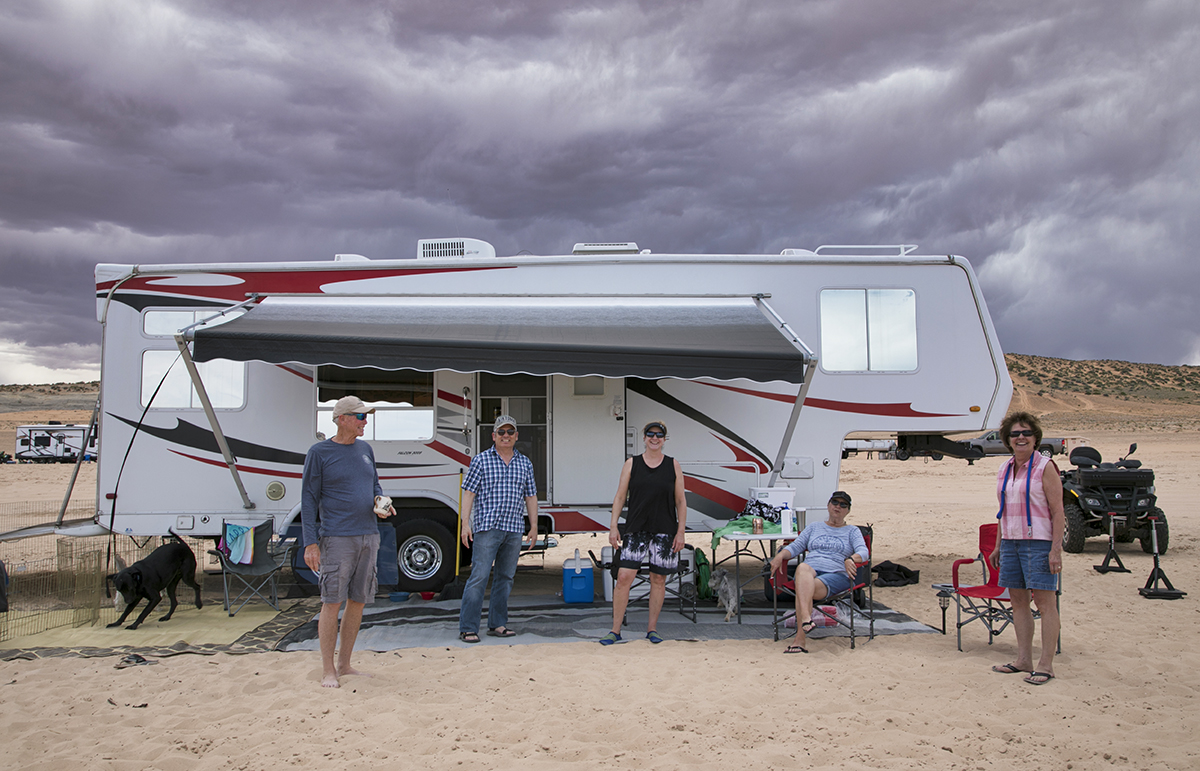 Campers stand in front of an RV in a sandy terrain