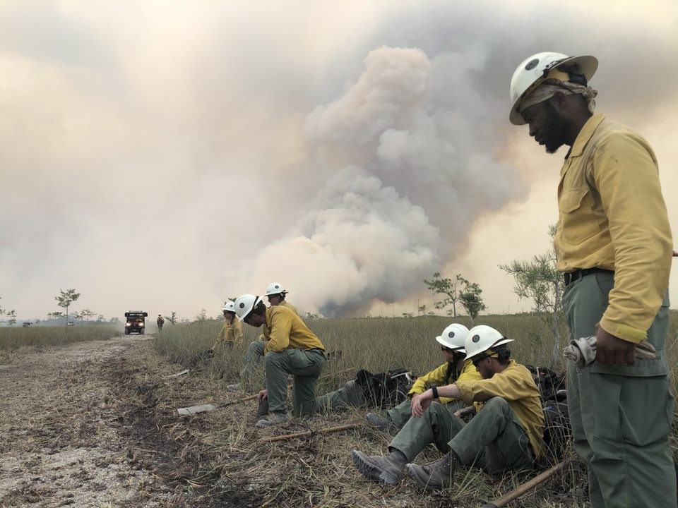 A group of firefighters stand in the foreground as they suppress a wildfire. They wear yellow shirts and green pants, and three of them are sitting down to rest before going back to work. Shelby Fox, BLM photo.