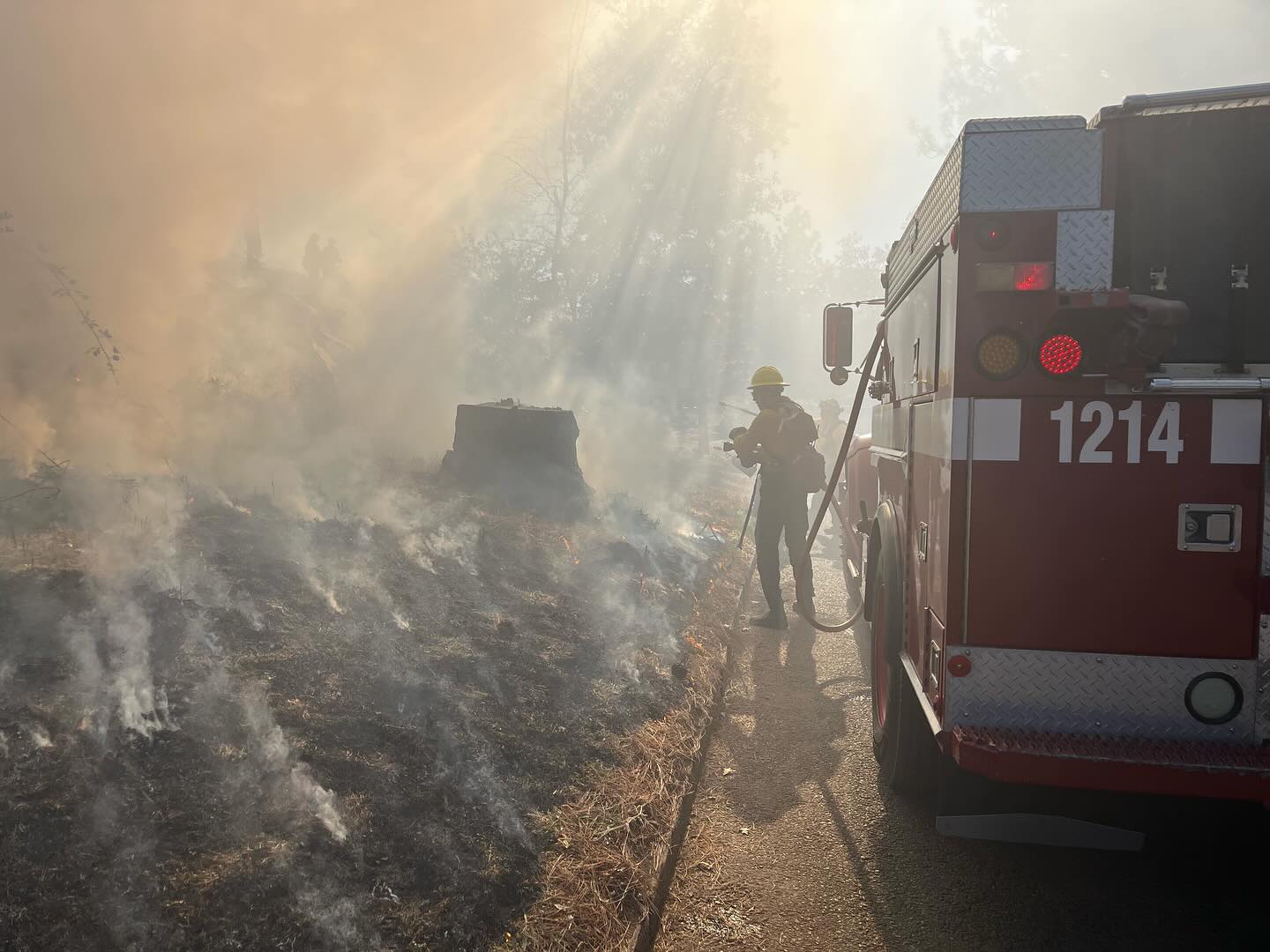 A firefighter sprays water on a wildfire as he walks next to a red fire engine. 