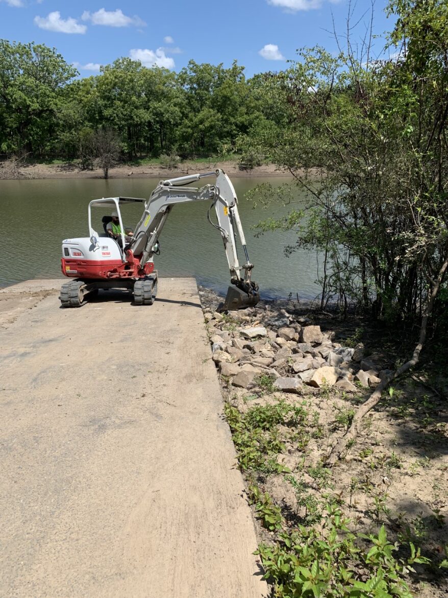 Excavator sitting on a concrete boat launch moves large rocks surrounding the launch.