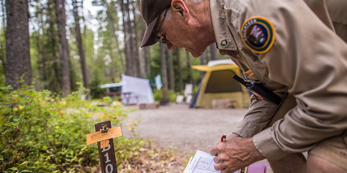 Man in volunteer uniform crouches near campground marker. 