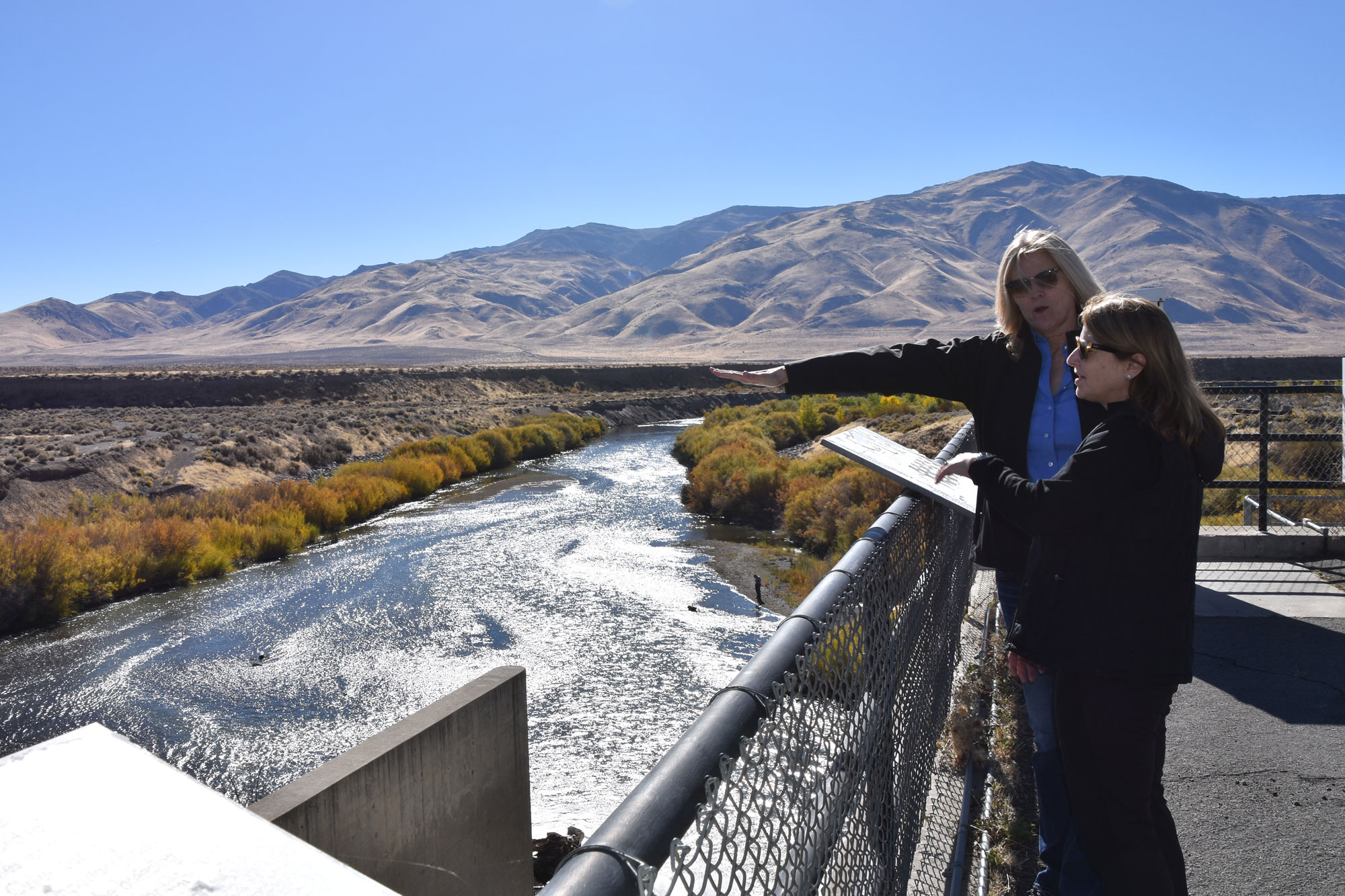 Two people standing on platform above river. Fall colors and mountains in the backdrop. 