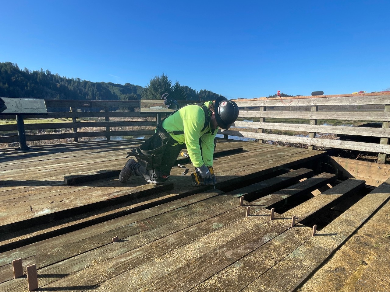 A construction worker removes a deteriorating wooden board on a brown observation deck.