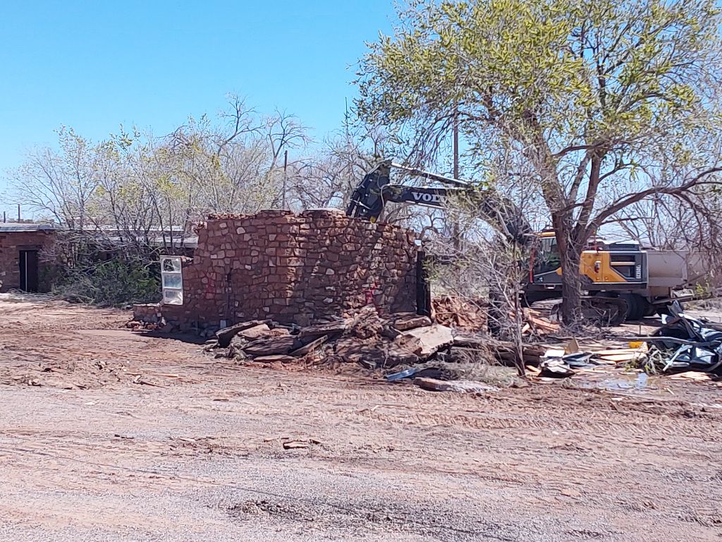 Black and yellow construction vehicle demolishes red brick building in construction area.