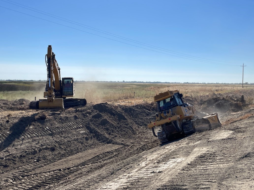 Two yellow construction vehicles sloping dirt along a waterway.