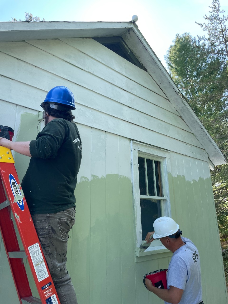 Two maintenance workers paint a white wooden facility green.