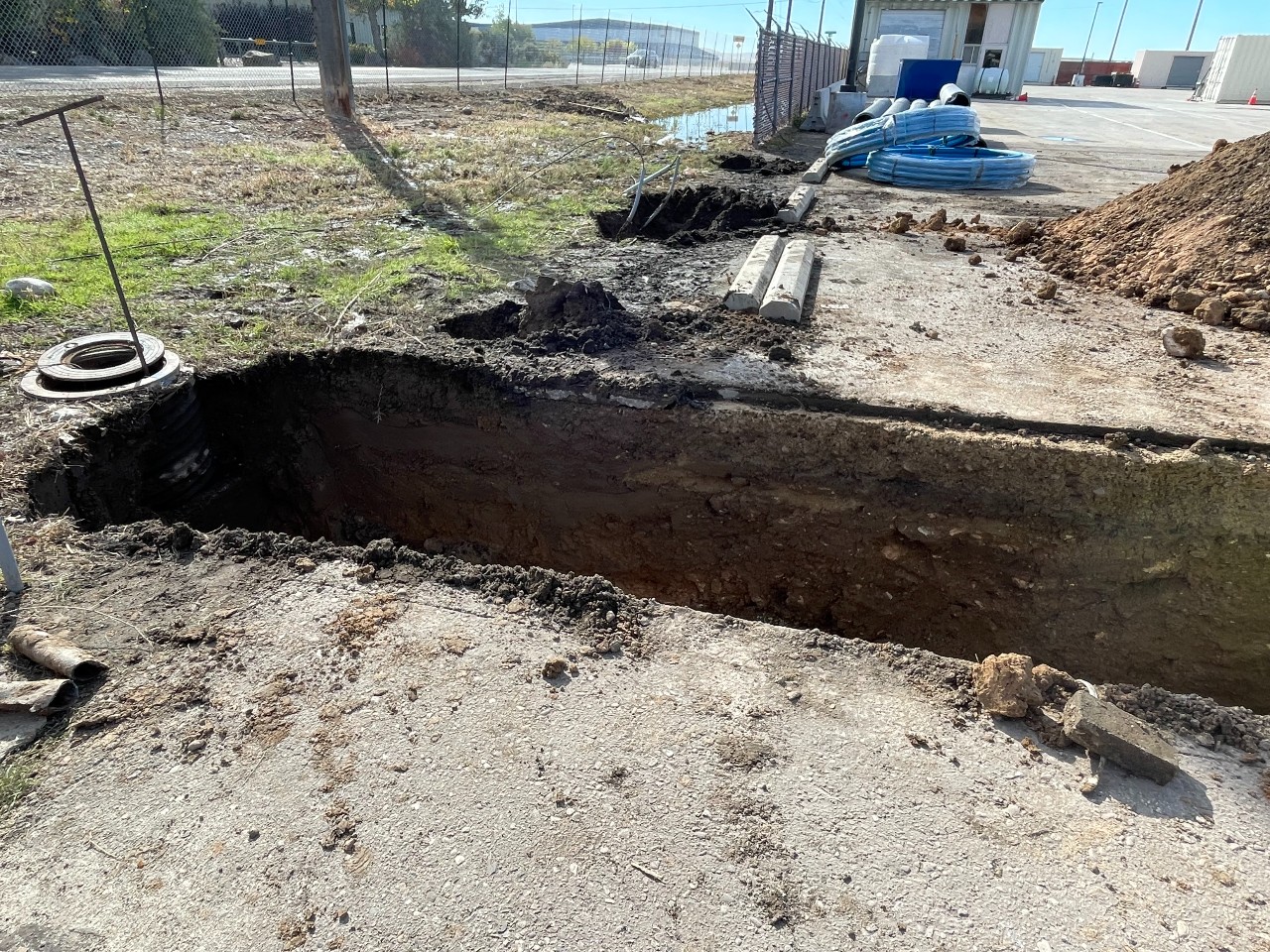 A dirt ditch surrounded by construction materials with gray fencing in the background.