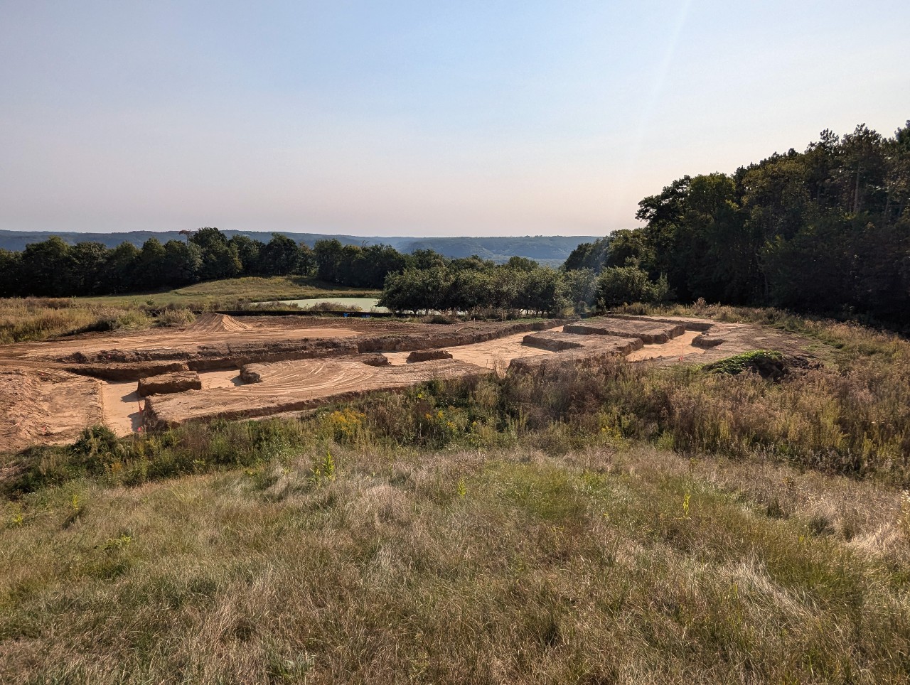 Construction site with brown dirt piles surrounded by grass and dark green trees in the background.