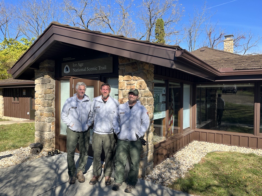 Photograph of three people standing in front of building with Ice Age National Scenic Trail sign on the exterior.