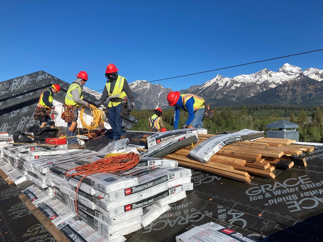 Construction workers stand next to construction materials on roof with white mountains in the background.