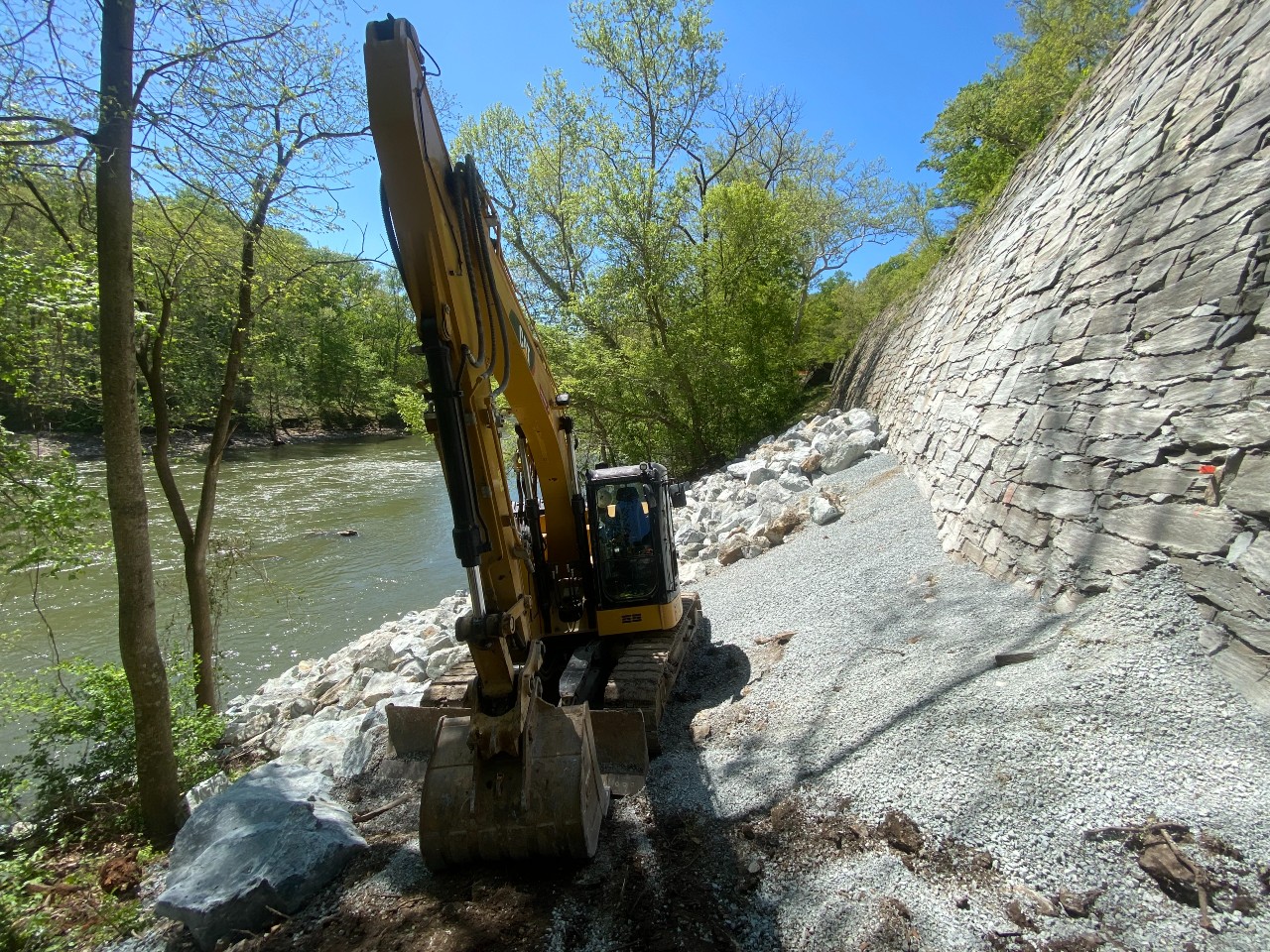 Yellow construction vehicle sites next to gray dry stone retaining wall next to river and greenery.