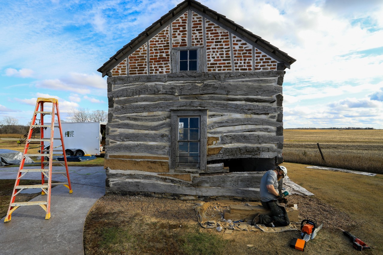 Man wearing hard hat, gloves, and chaps performs historic cabin restoration work.