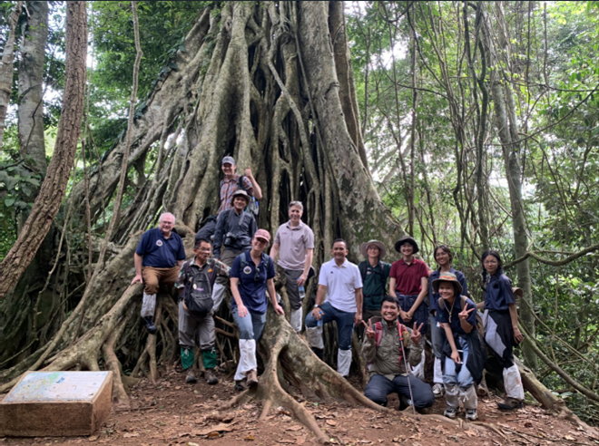 a group poses in front of a large tree with many roots in SE Asia