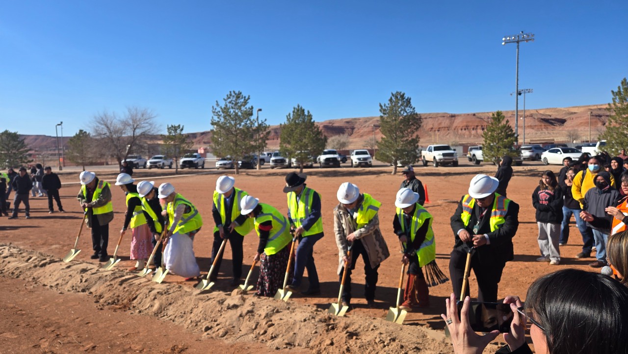 Group of people in yellow construction vests and hard hats use shovels to break ground on dirt plot.