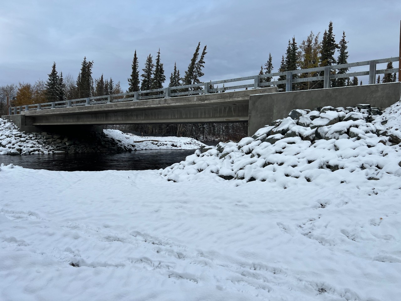 Newly built concrete bridge crosses over dark river surrounded by snow.