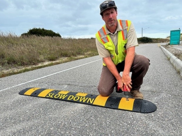 Man in yellow construction vest drills yellow and black speed bump on paved road.