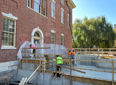 Construction workers stand on concrete platform with wooden scaffolding attached to the exterior of brick building.