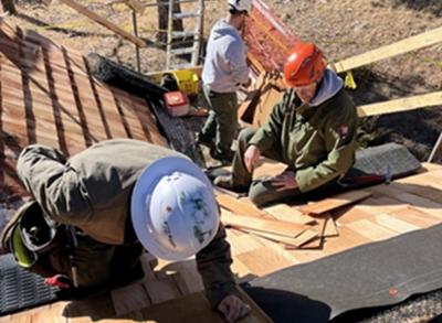 Three construction workers in hard hats place wood beams on the side of a historic cabin.