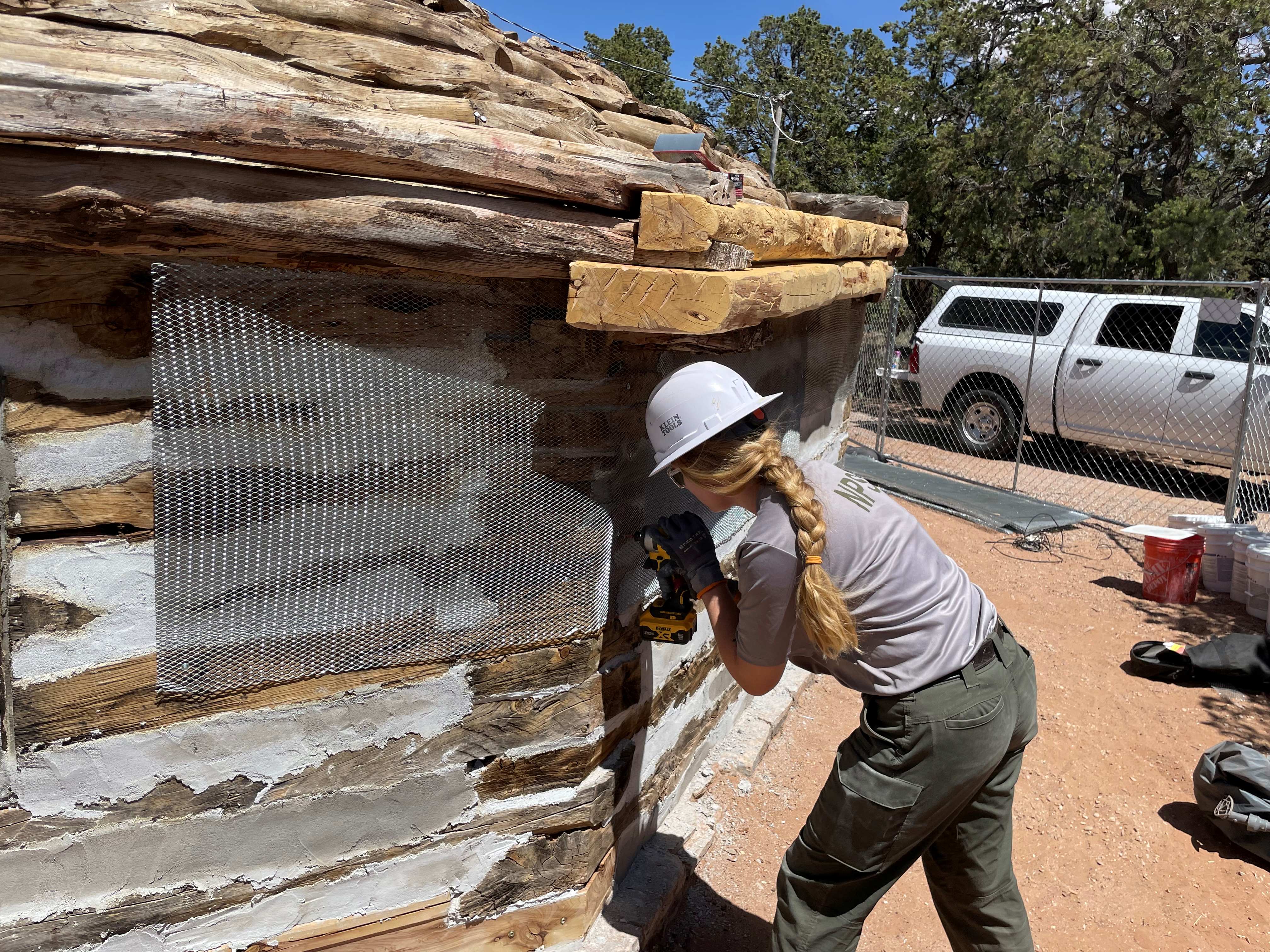 Woman in white hard hat drills exterior of historic wooden housing structure.