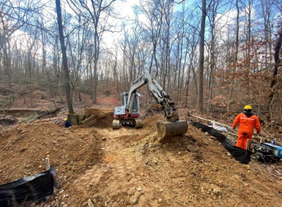 An excavator pulls up dirt in wooded area surrounded by temporary fencing.