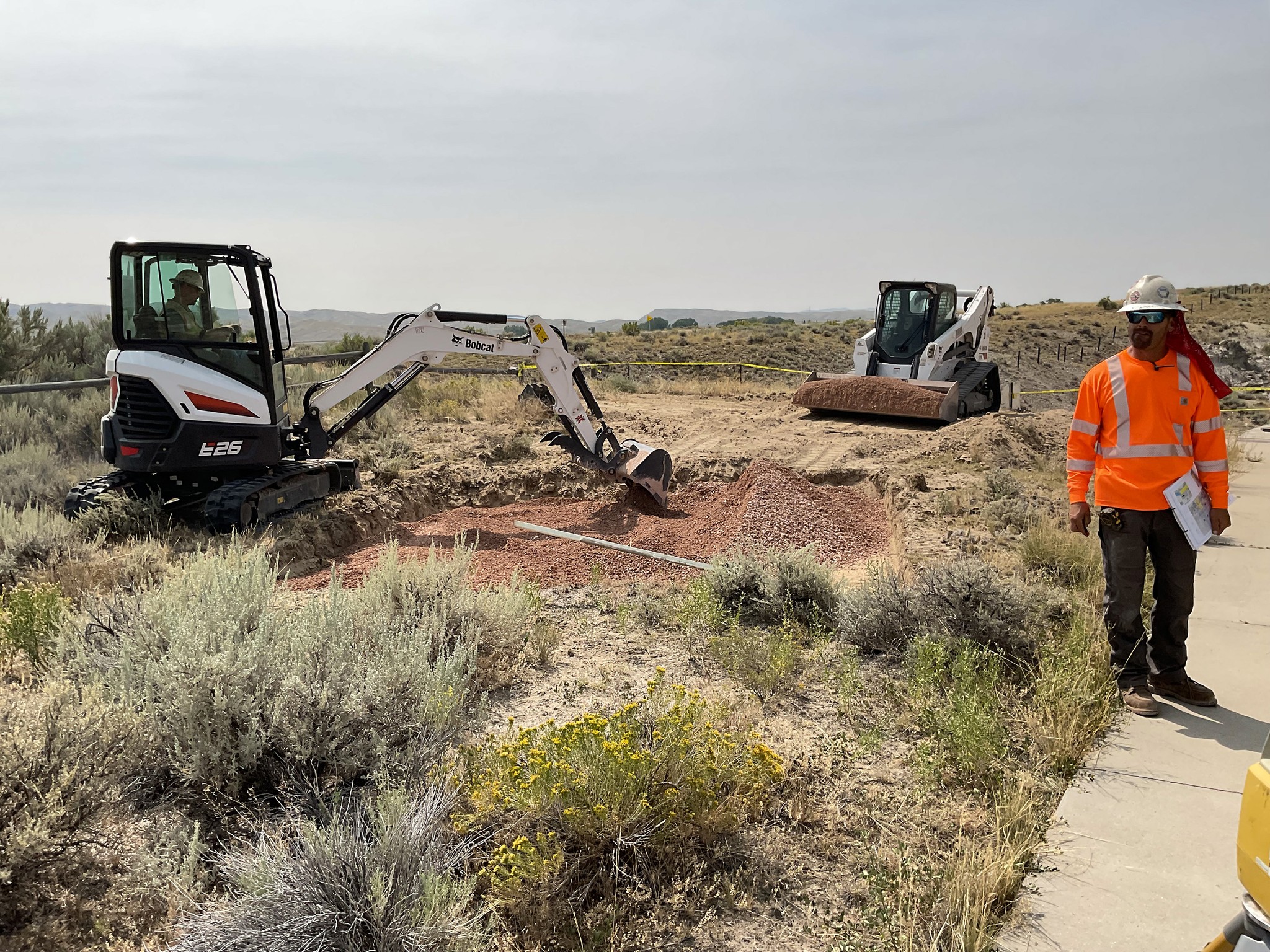 Man in an orange construction shirt and hard hat stands to the side of construction machinery moving dirt.