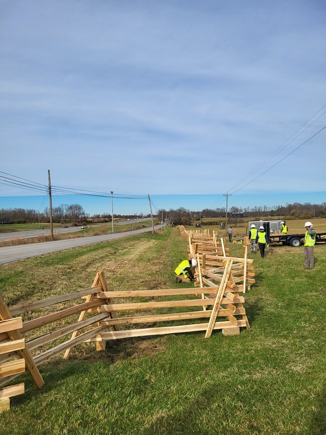 Construction workers in yellow vests repair wooden fence that runs alongside paved road.