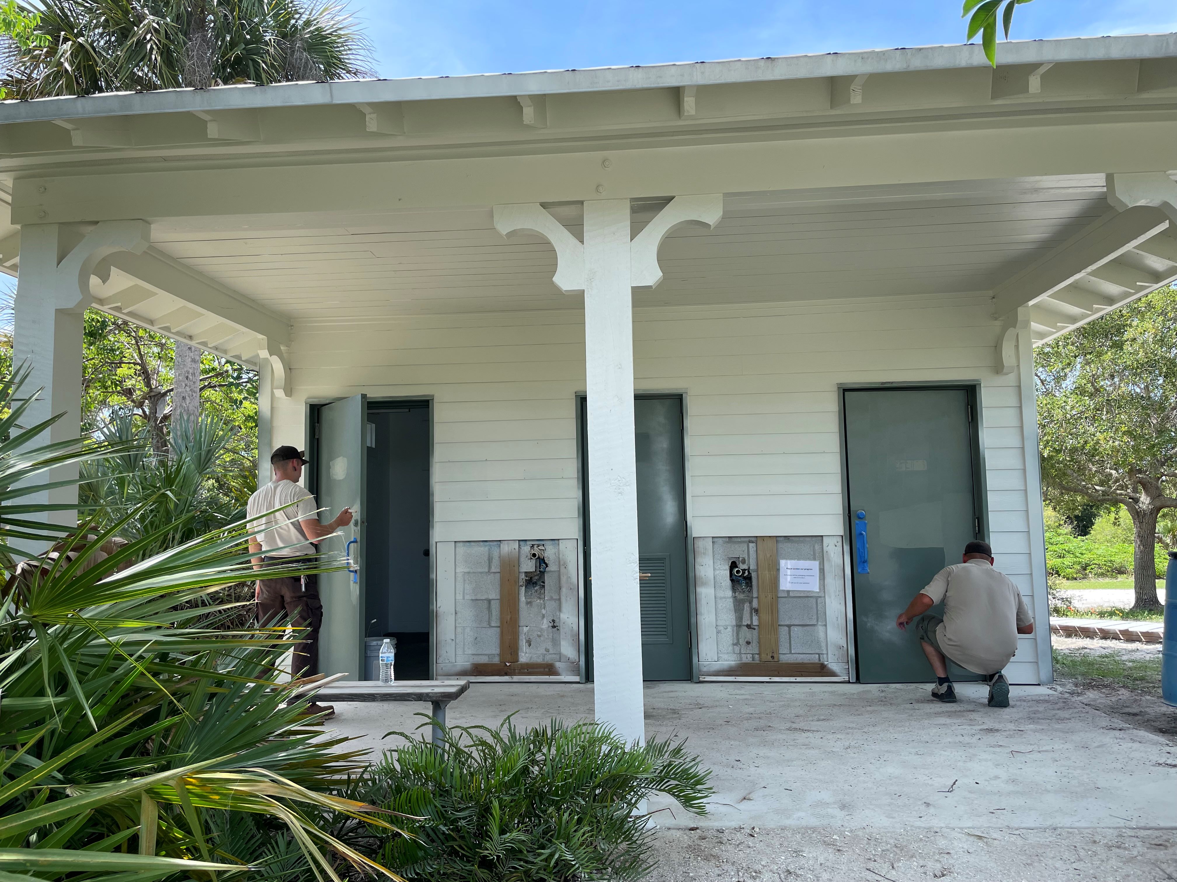 Two men stand on concrete porch to paint the exterior of white comfort station with green doors.