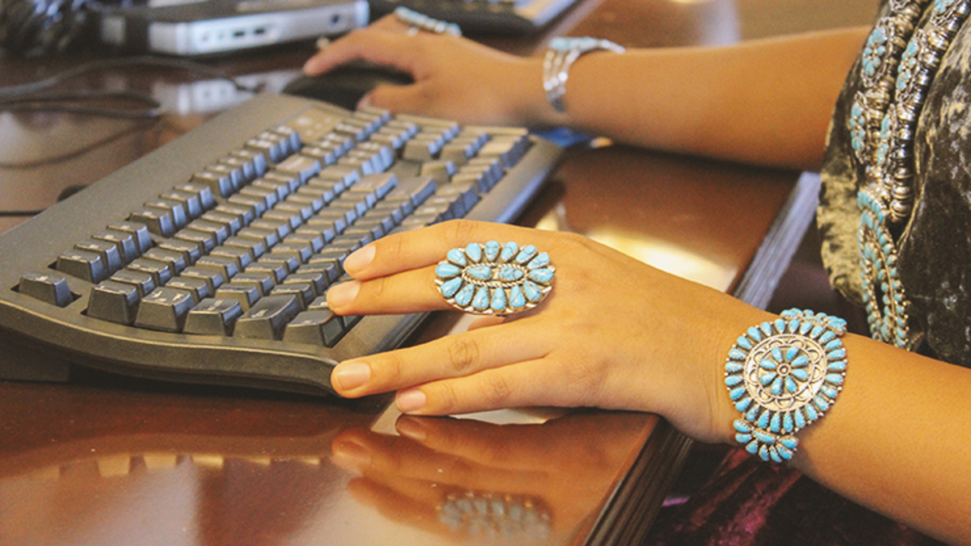 A view of a person's hands with turquoise jewelry typing at a keyboard and using a computer mouse.
