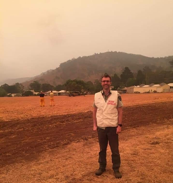Peter stands in a field with a smoke-filled background. 