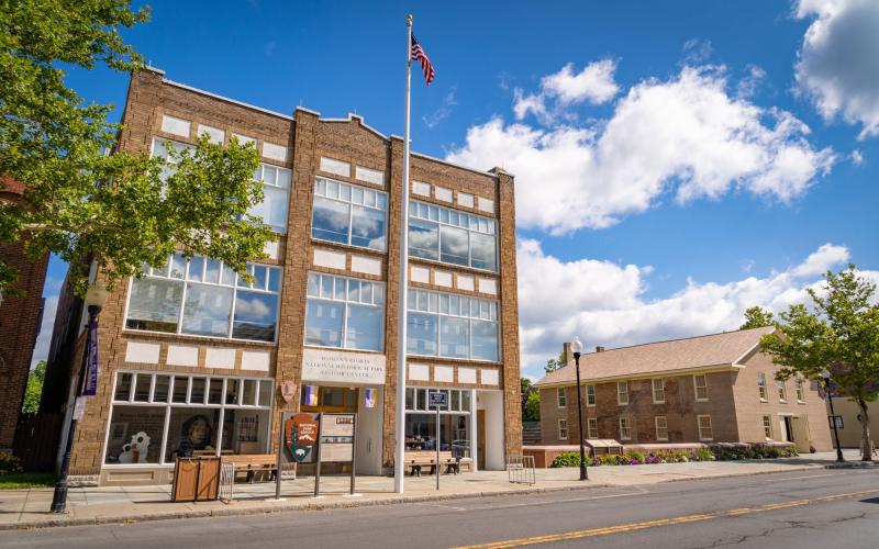 A three-story stone building with large glass windows and an American flag sits close to the road, and to its right, the brown brick Wesleyan Chapel is visible.