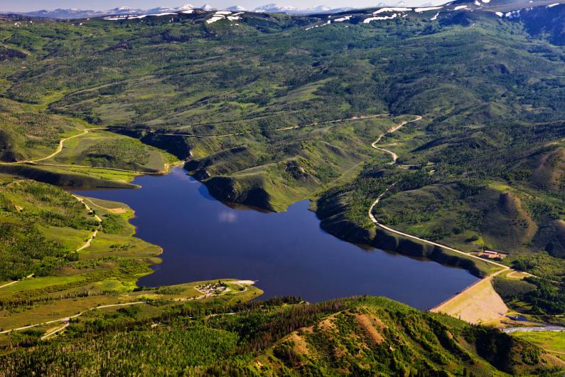 Aerial view of Currant Creek Dam Reservoir with snow capped Uinta Mountains in the background.