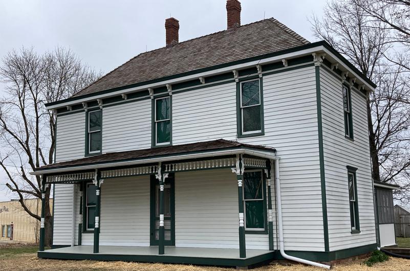 White house with green trim sits atop a lawn with trees on either side under a gray sky.