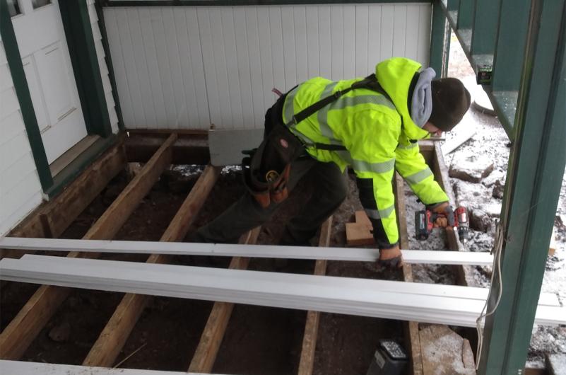 Construction worker crouching to drill a hole in wooden planks laying on the porch of a house.