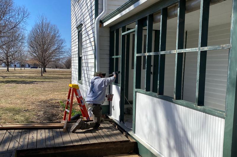 Man painting the side of a white house with green trim, next to small orange ladder.