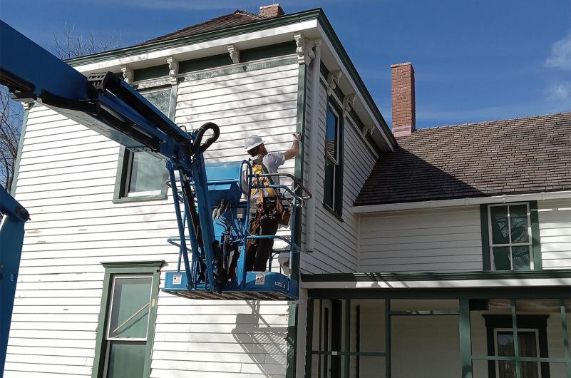 Man in a hard hat stands on an aerial work platform to paint the green trim of a white house.