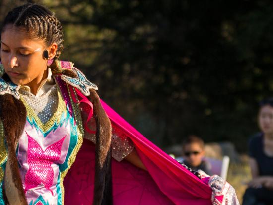 A young woman in traditional tribal clothing dances outside as a small group of people look on.