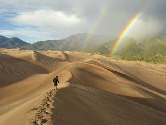A hiker on the ridge of a sea of dunes with a double rainbow and mountains in the distance