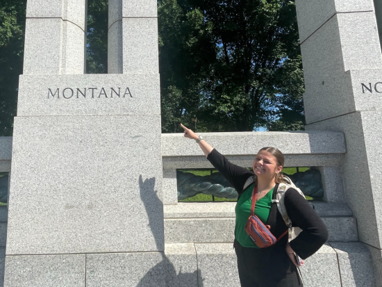 Zoe Belinda, Office of Wildland Fire intern, standing next to the Montana block of the World War II Memorial in Washington D.C. She points to the word "Montana."