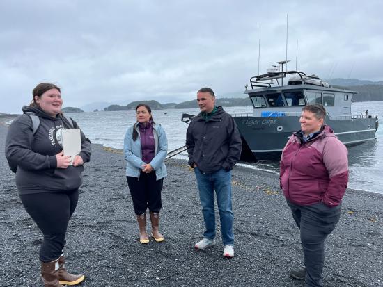 Four people stand along gravel beach in front of boat. 