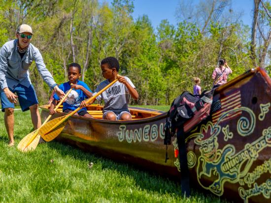 Two kids in a canoe on land, an adult teaches them to paddle.  