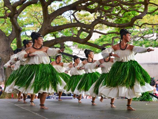 Nine dancers are dancing on stage wearing white dresses and grass skirts.