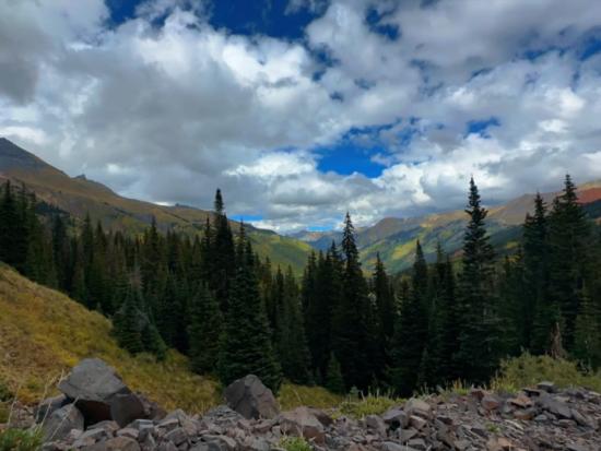 Rocks, grass, and evergreen trees are seen across a mountainous landscape under a cloudy blue sky