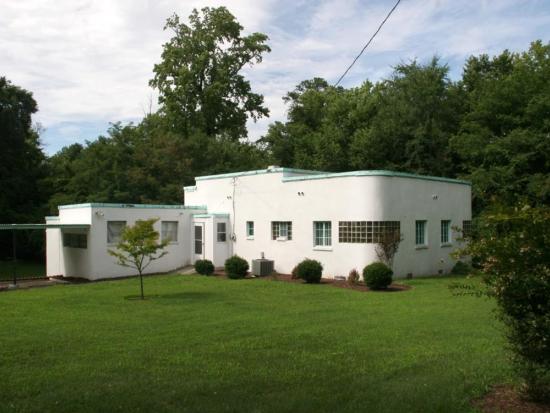 A white building on green grass in front of green trees and a blue sky with white clouds. South elevation of garage (at left) and west and south elevation of house (at right) showing carport and garage (enclosed and altered into a studio by Meredith), kitchen entrance and windows of utility room and bathroom at center, and bedrooms with block glass curved corners to right. 