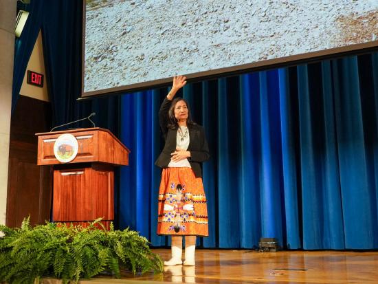 Secretary Haaland on a stage next to a podium waving to a crowd.