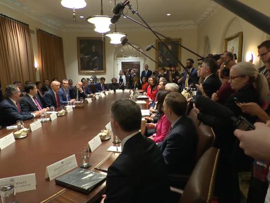 President Trump and his Cabinet sit at a large brown conference table in front of a large gathering of media members