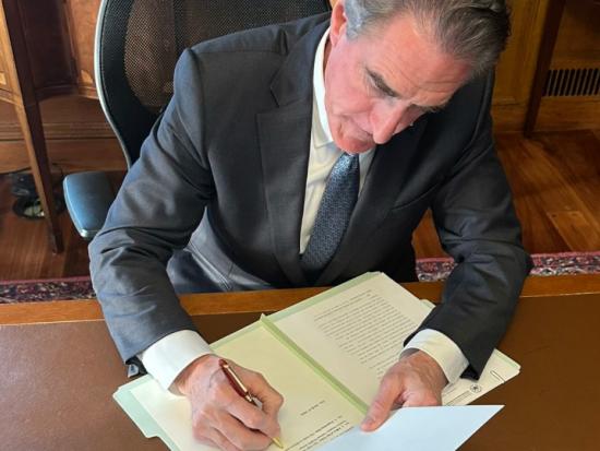 Secretary Burgum sits at a brown desk with pen in hand to sign a document 