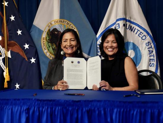 Two women hold up signed document with Department of Labor, Department of the Interior and EPA flags in the backdrop
