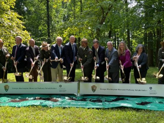 Various people, wearing suits and green uniforms, dig dirt behind a banner.