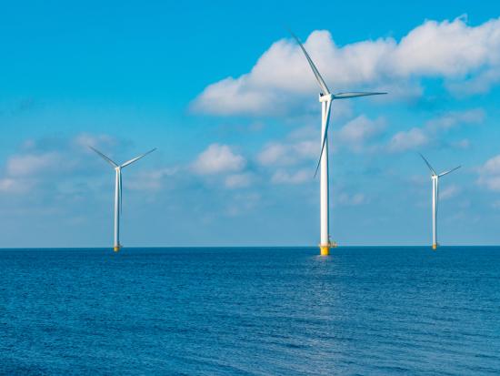 Three wind turbines stand in the ocean with a blue sky and white clouds in the background.